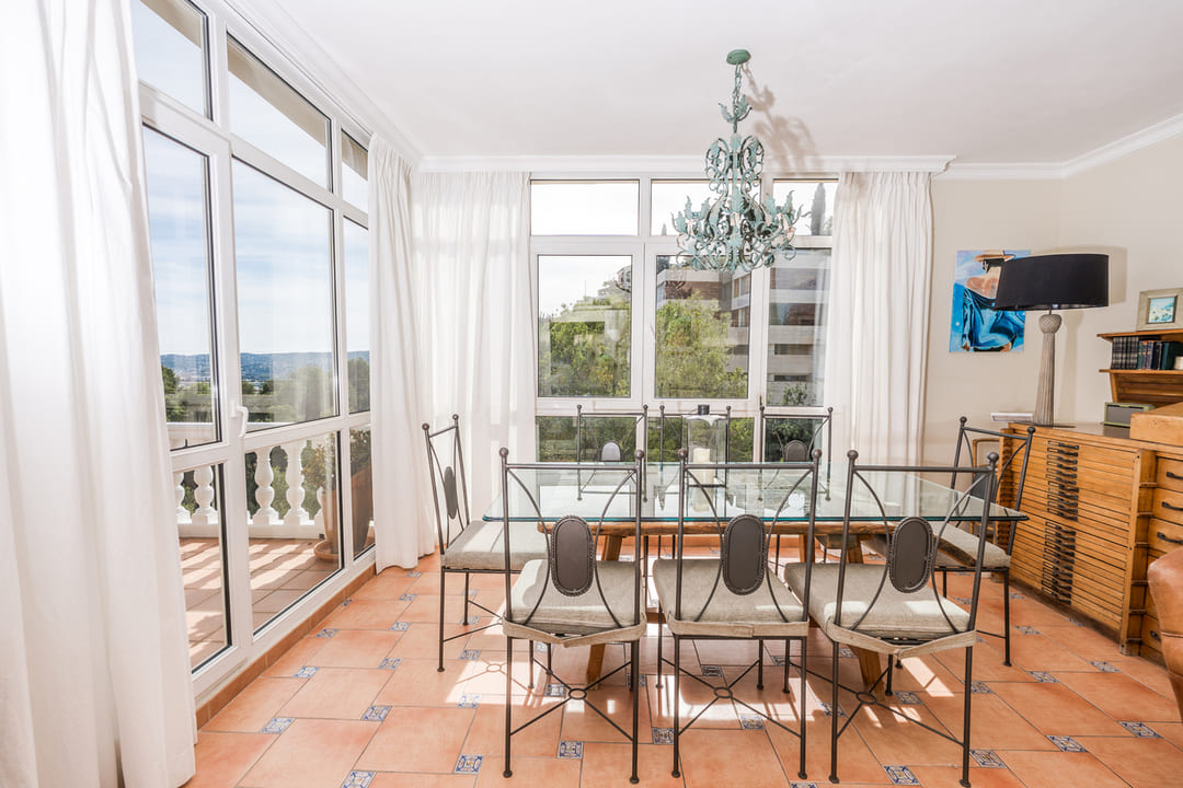 Comedor con mesa de cristal y sillas de forja sobre suelo de terracota. Grandes ventanales inundan el espacio de luz natural y ofrecen vistas al entorno exterior.