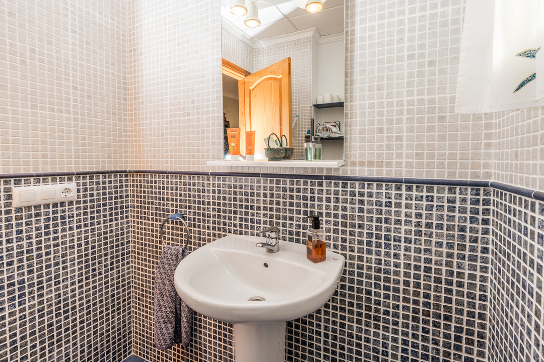 Cuarto de baño con lavabo de pedestal blanco, paredes de mosaico en tonos blancos y azules, y un espejo con luminaria integrada y repisa.
