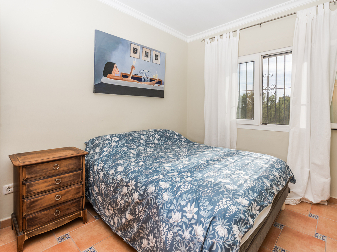 Dormitorio con cama doble, edredón de motivos florales azules, mesita de noche de madera y suelos de baldosa con detalles decorativos.