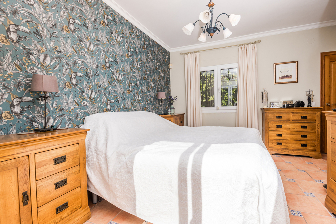 Dormitorio con pared de acento de papel tapiz tropical, cómodas de madera maciza y suelo de baldosas de terracota con detalles decorativos. Incluye iluminación clásica.
