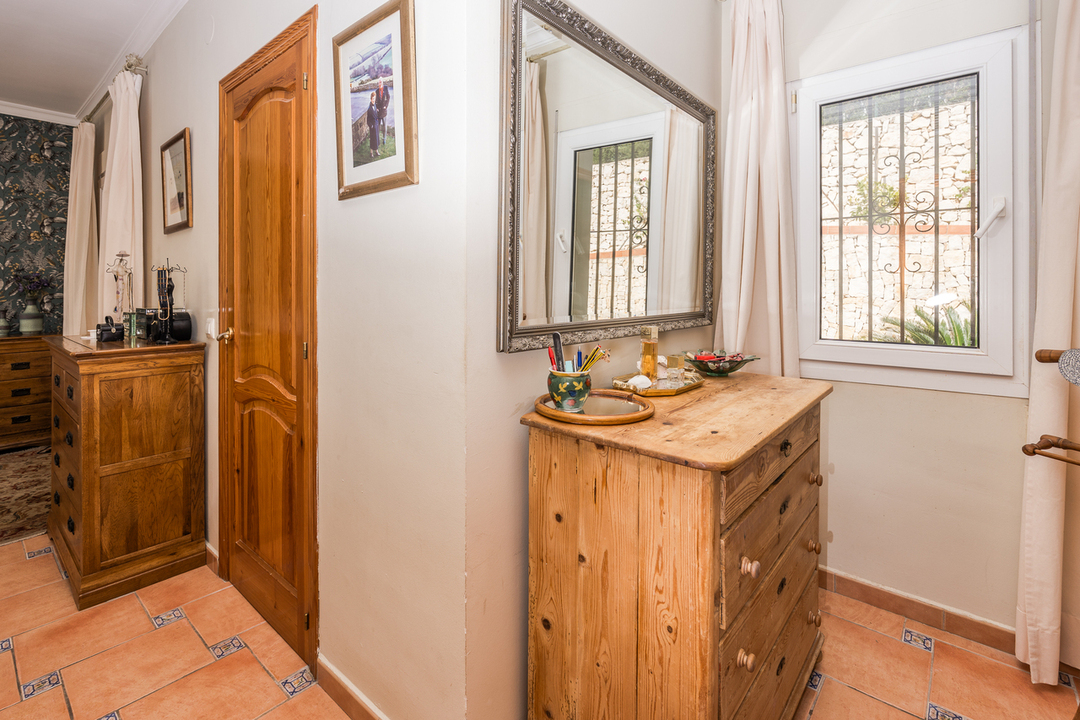 Rincón de dormitorio con cómoda de madera, suelos de terracota con detalles cerámicos y un gran espejo de marco plateado junto a la ventana con rejas.