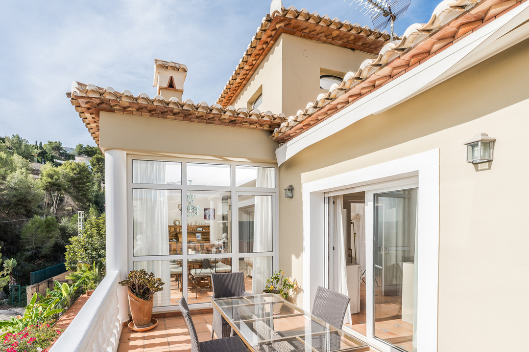 Terraza con suelo de terracota, comedor exterior de cristal y balaustrada blanca. Se aprecia un cerramiento acristalado y vistas a la arquitectura mediterránea y la montaña.