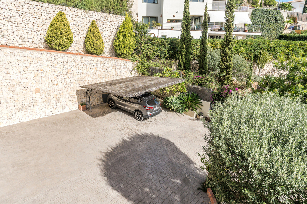 Zona de aparcamiento exterior con suelo adoquinado y pérgola de brezo, delimitada por un muro de piedra seca y vegetación mediterránea cuidada.