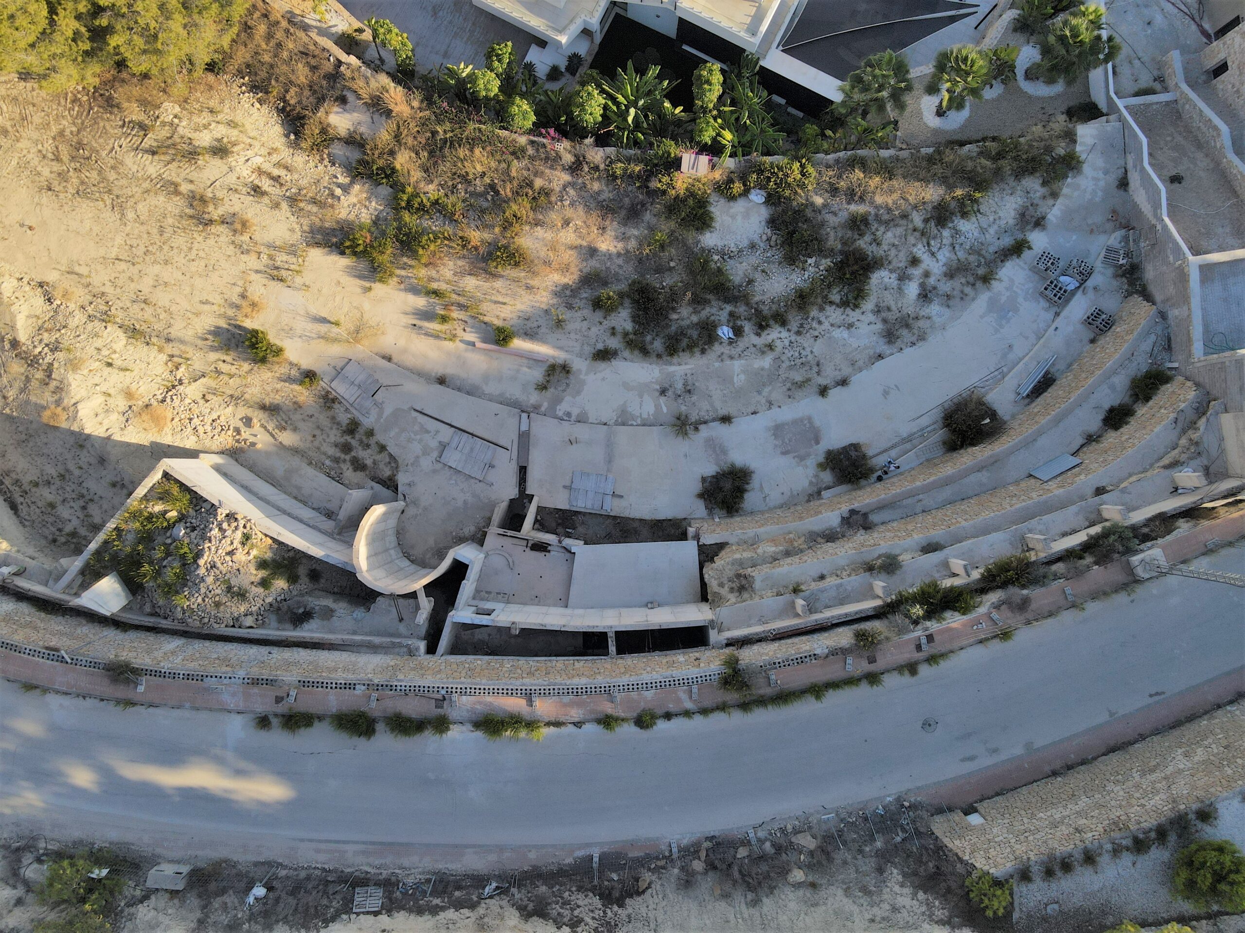 Vista aérea de una parcela en construcción con cimientos curvos de hormigón y muros de contención de piedra integrados en el terreno inclinado.