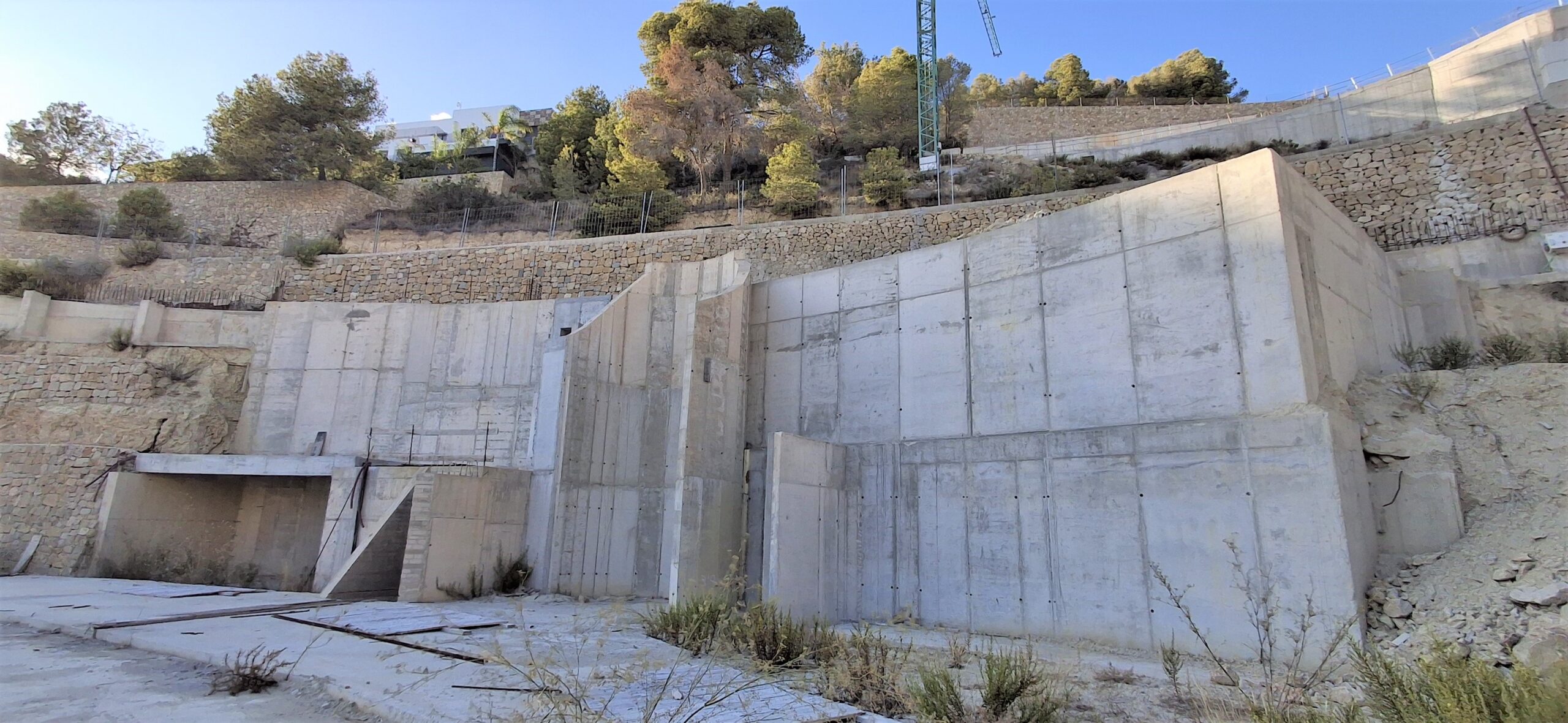 Vista de los muros de contención de hormigón armado y bancales de piedra integrados en una parcela inclinada con vegetación mediterránea.