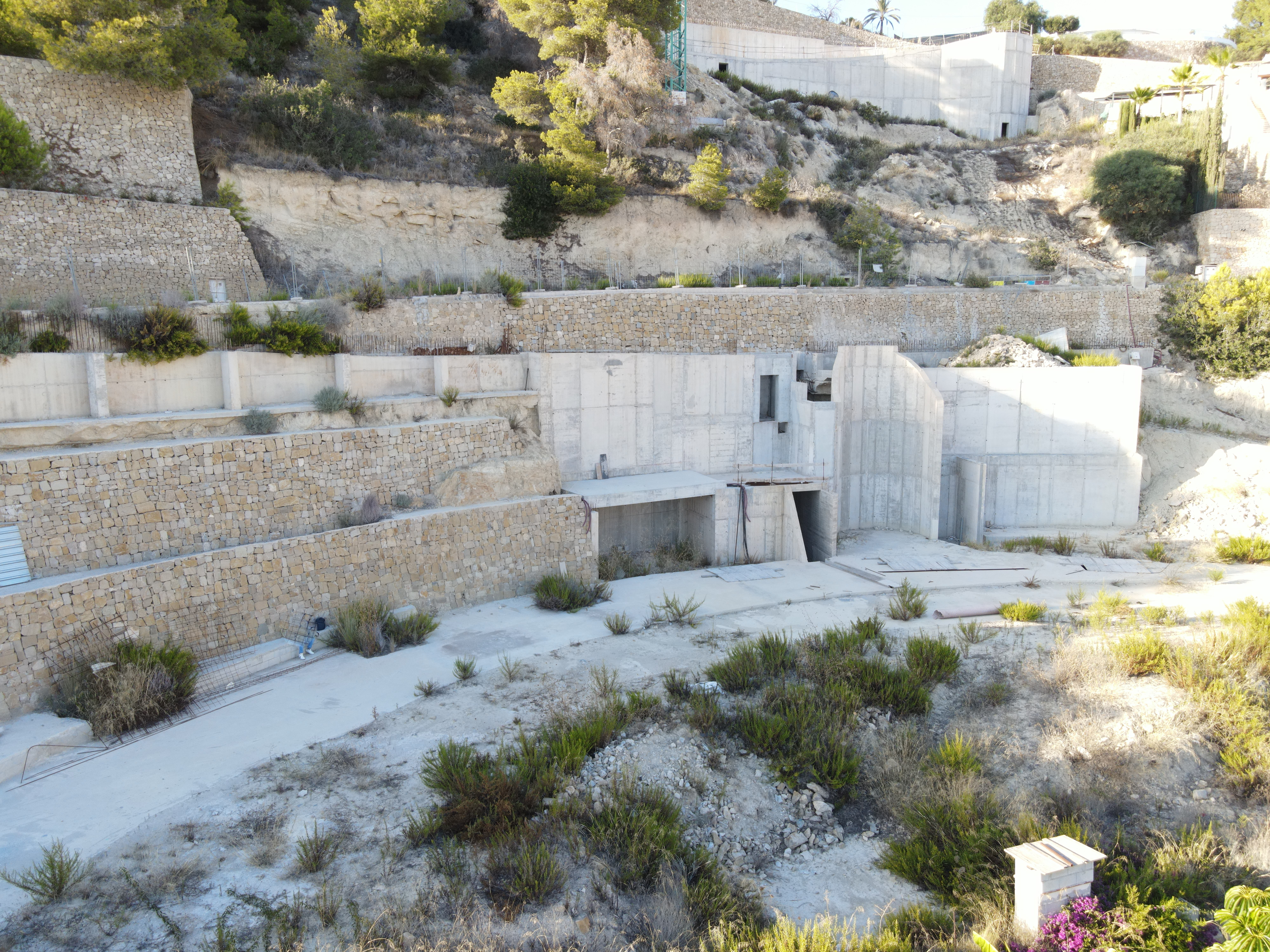 Vista de obra con muros de contención de piedra seca escalonados y estructuras de hormigón armado integradas en la ladera con vegetación.