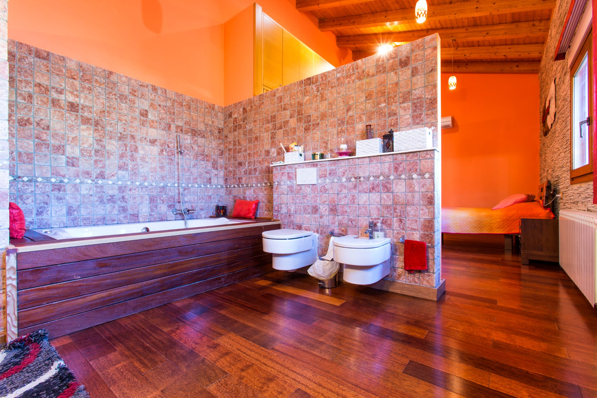 Baño integrado con bañera revestida en madera, paredes de mosaico rústico y sanitarios suspendidos, bajo un techo de vigas de madera y suelos de parqué oscuro.