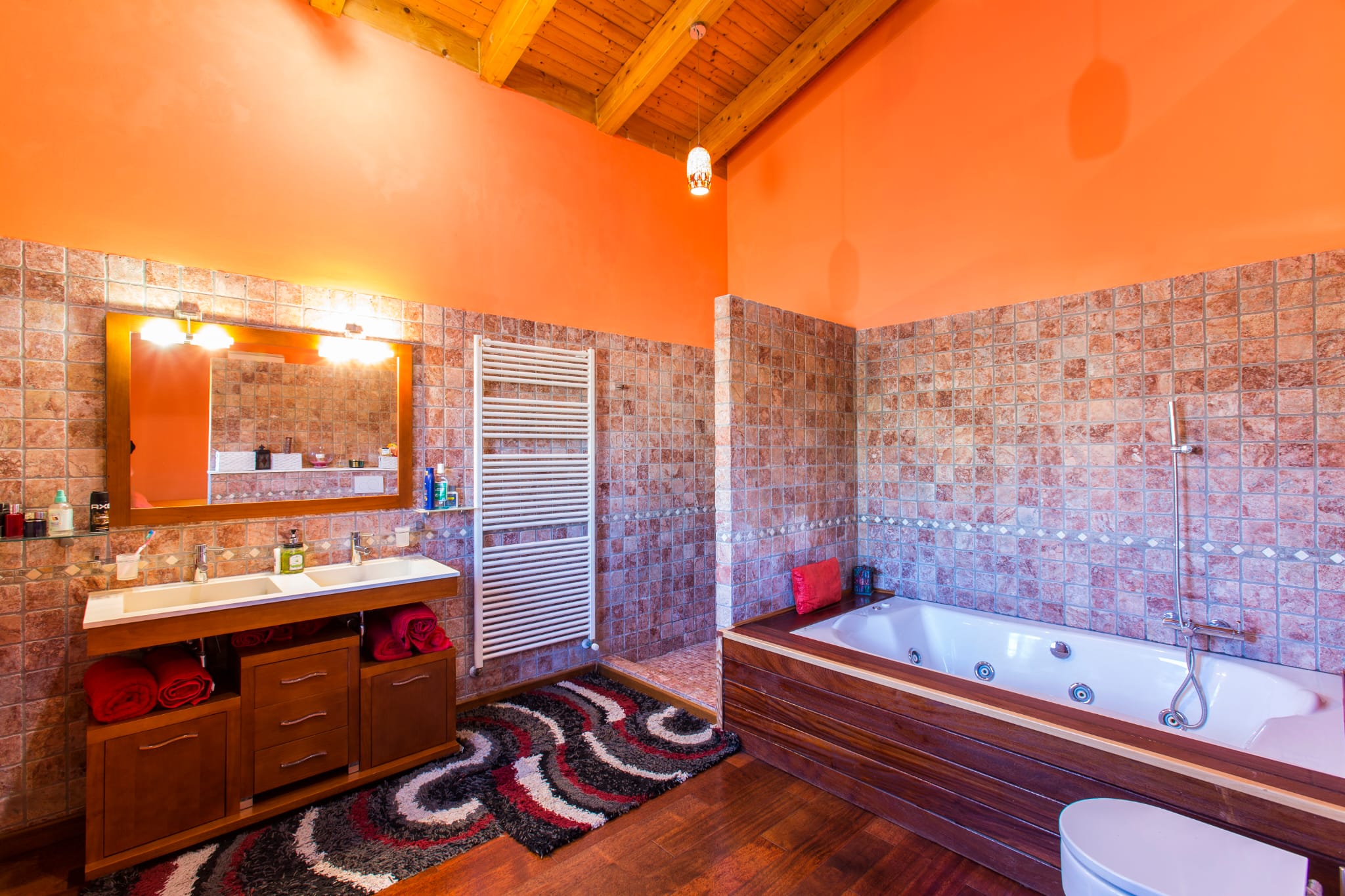 Baño rústico con techos de vigas de madera, lavabo doble sobre mueble de madera oscura, bañera de hidromasaje revestida y paredes en tonos terracota y naranja.