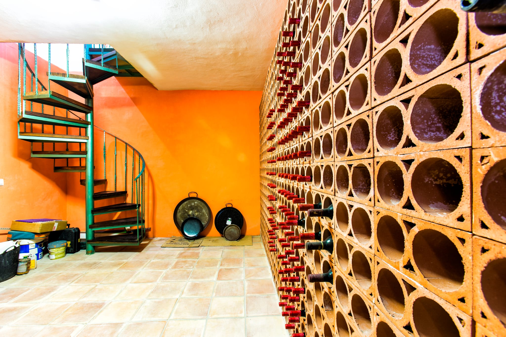 Bodega con botellero de terracota en nido de abeja, pared naranja y una escalera de caracol de metal verde con peldaños de madera oscura.