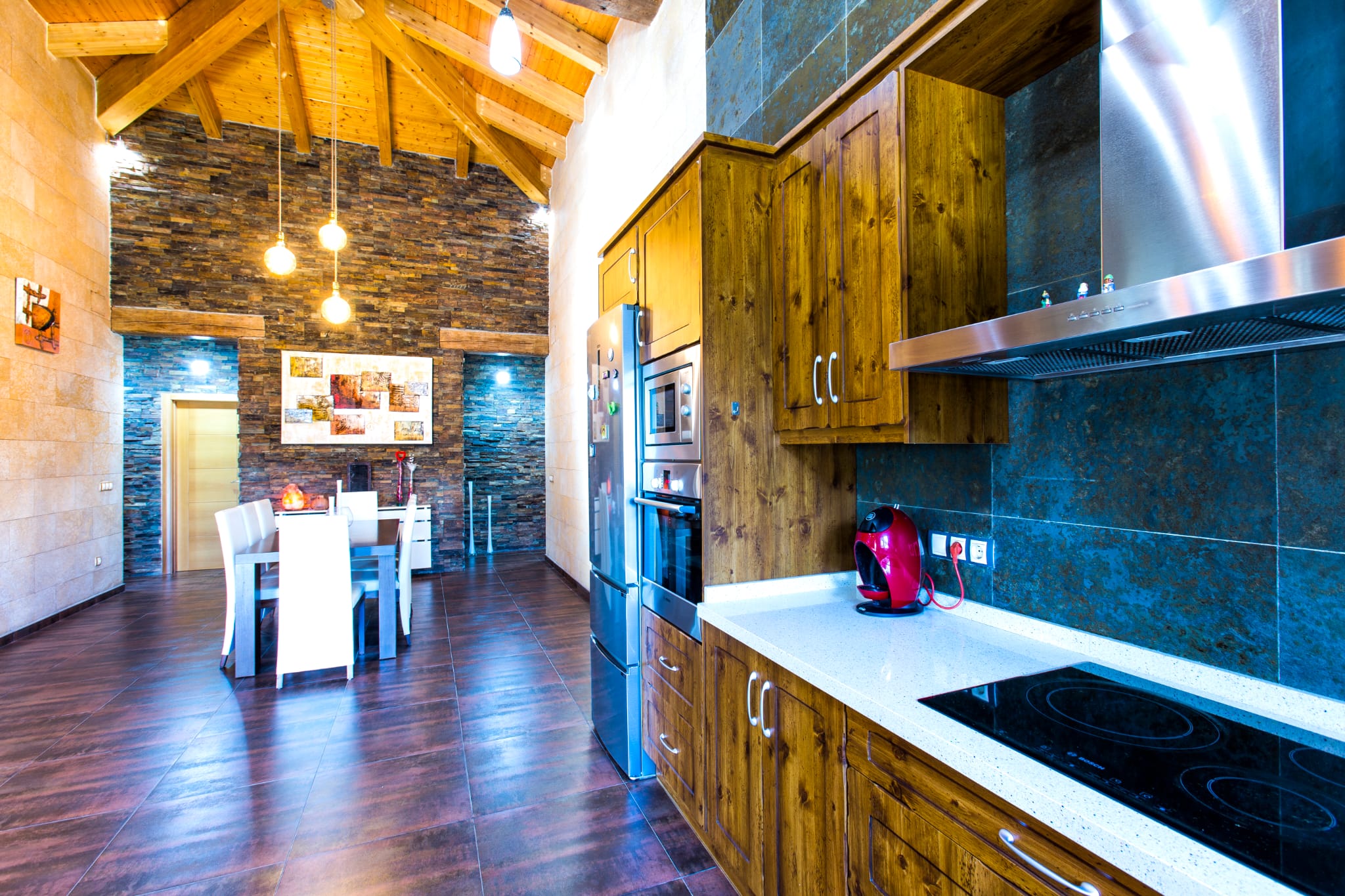 Cocina con armarios de madera rústica, encimera blanca y electrodomésticos integrados, conectada a un comedor con paredes de piedra y techos de madera altos.