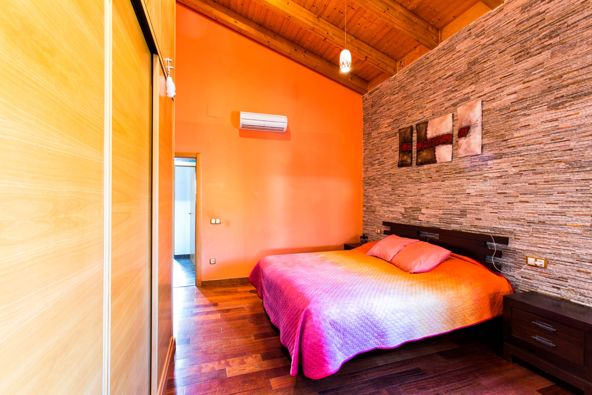 Dormitorio con techos de vigas de madera, pared de acento en piedra texturizada y armarios empotrados. Presenta suelos de madera, aire acondicionado y mobiliario oscuro.