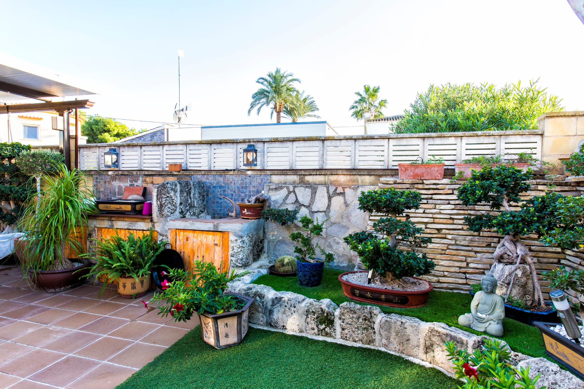 Jardín zen en patio con césped artificial, muros de piedra apilada y colección de bonsáis. Incluye encimera de piedra integrada con azulejos de mosaico azul.