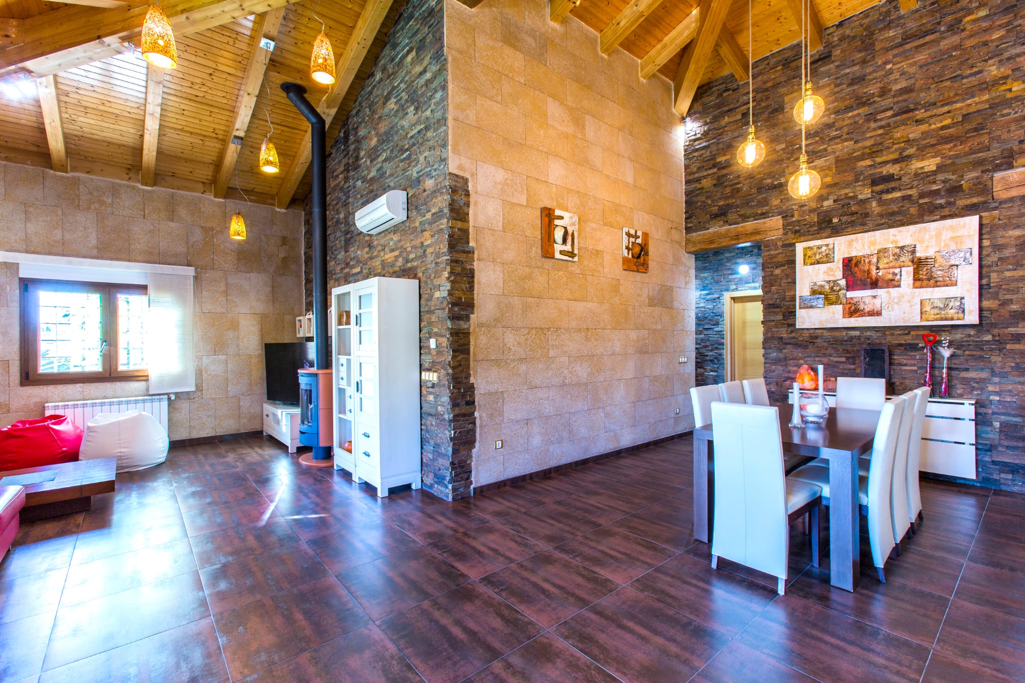 Salón comedor con techos de madera abovedados, paredes de piedra natural y una estufa de leña. Incluye mobiliario contemporáneo, lámparas colgantes y suelos cerámicos oscuros.