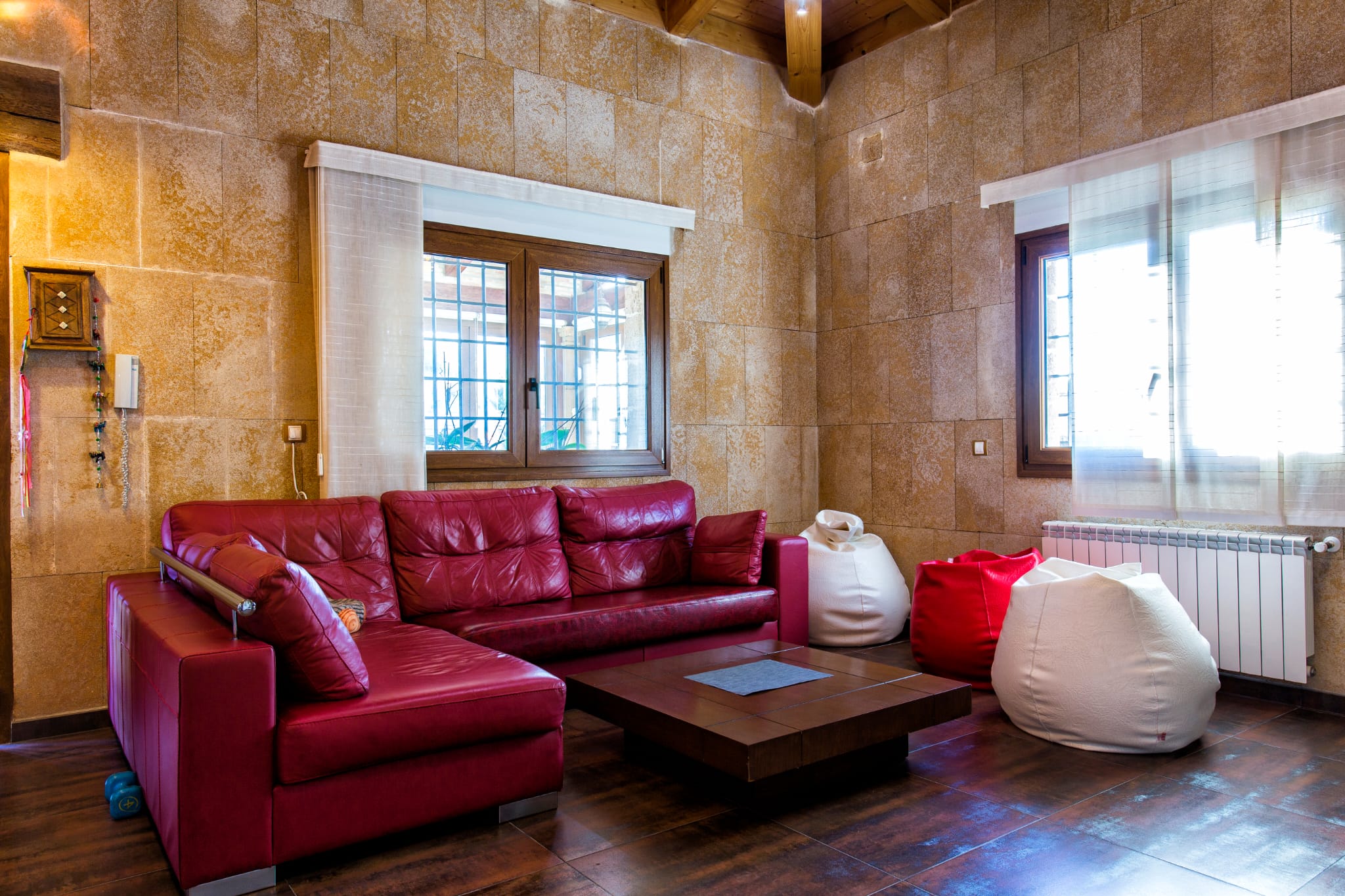Salón con sofá rinconera de cuero rojo, paredes revestidas de piedra natural y vigas de madera vistas. Incluye mesa de centro baja y pufs decorativos.