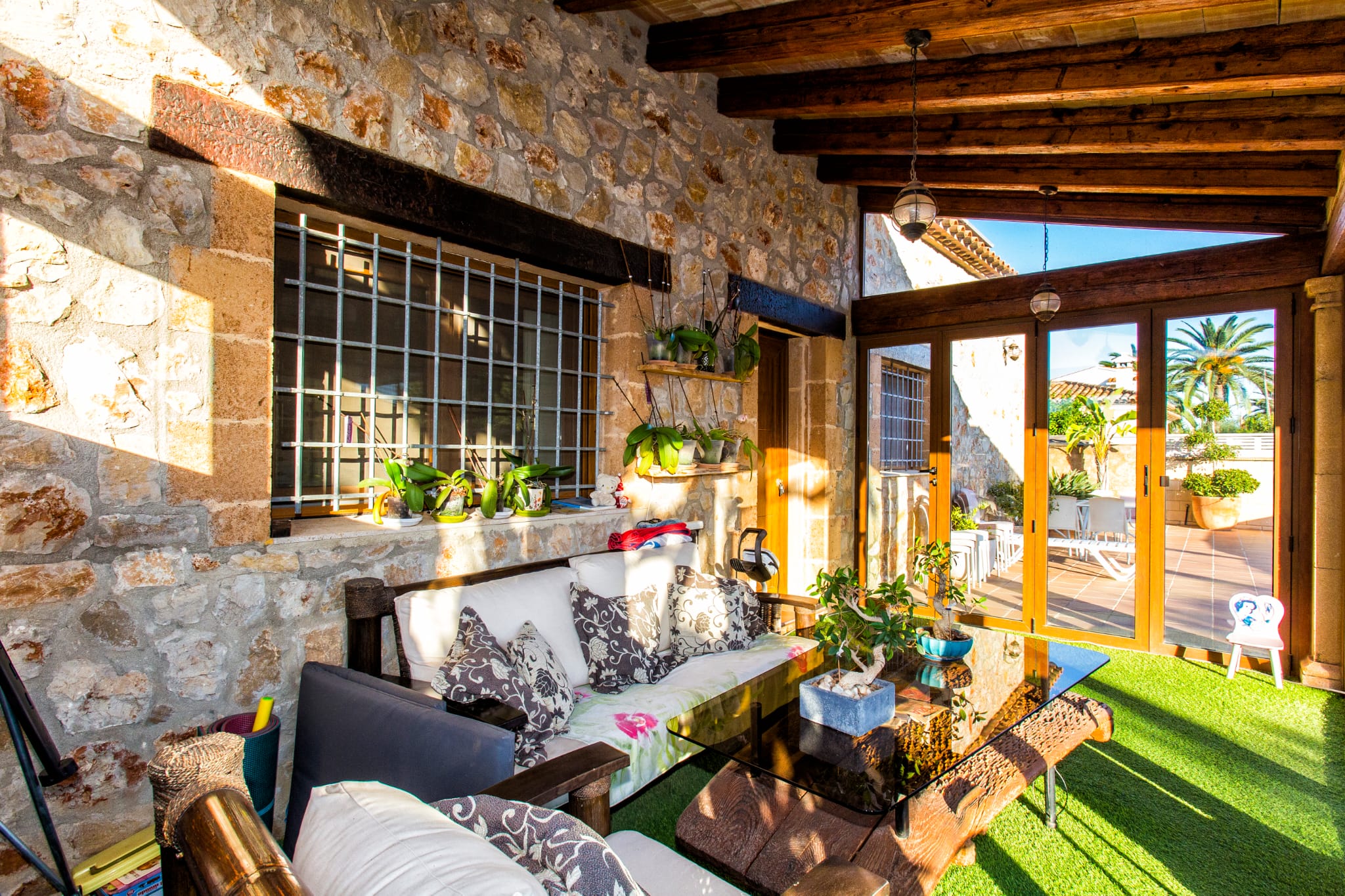 Terraza cubierta con paredes de piedra natural, vigas de madera oscura y zona de estar con mesa de cristal sobre césped artificial, abierta a un patio soleado.