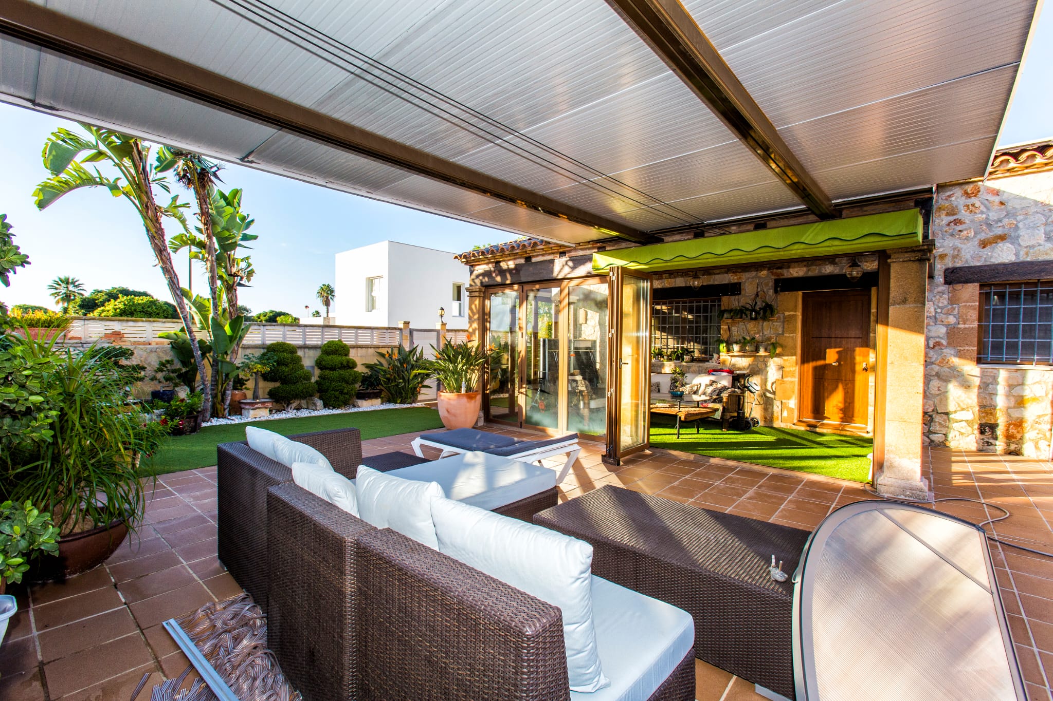 Terraza exterior techada con mobiliario de ratán sintético, suelo de baldosas cerámicas y jardín perimetral con césped artificial y vegetación tropical.