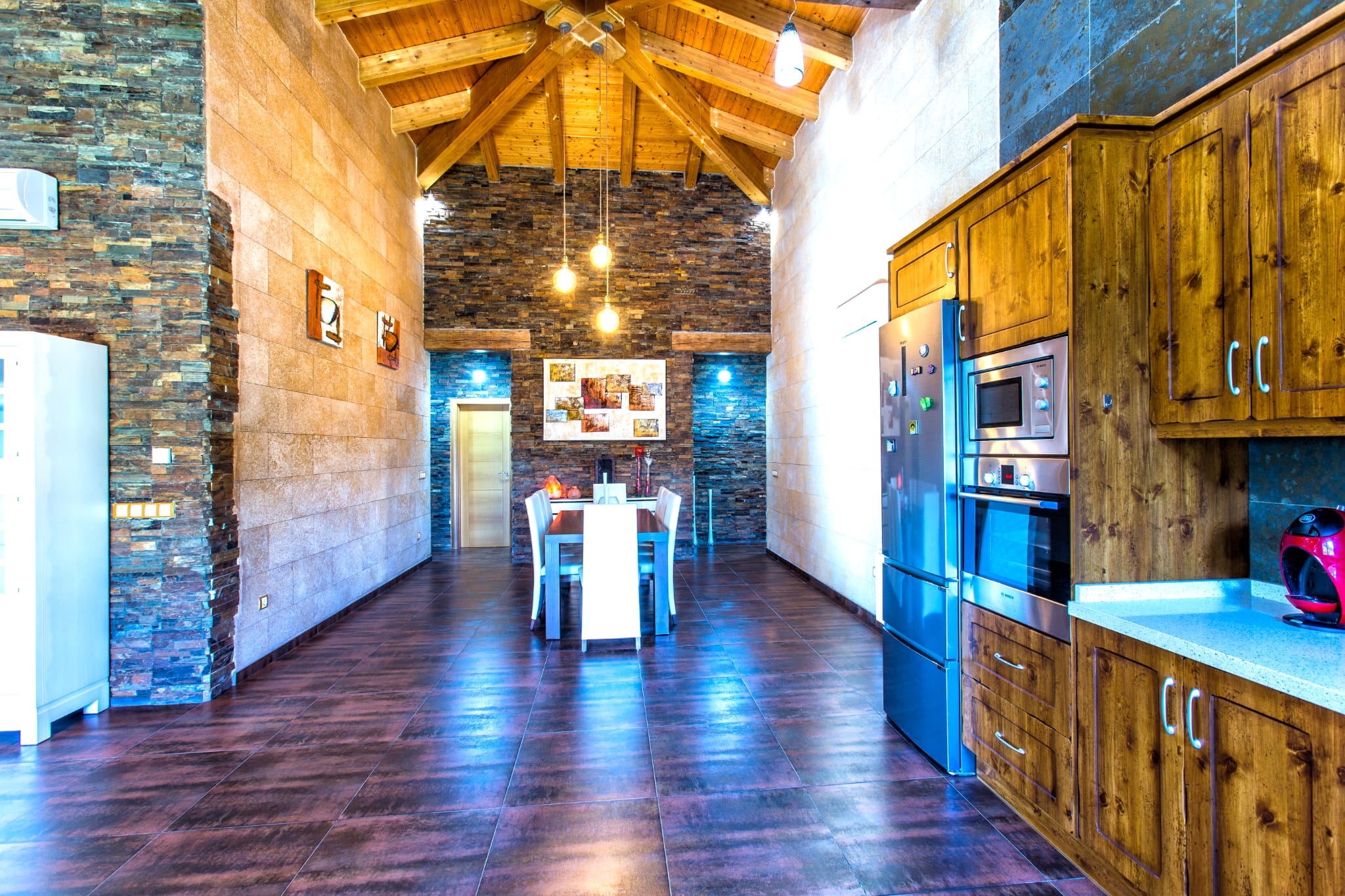 Zona de comedor y cocina con techos altos de madera, paredes de piedra natural apilada y electrodomésticos integrados de acero inoxidable.