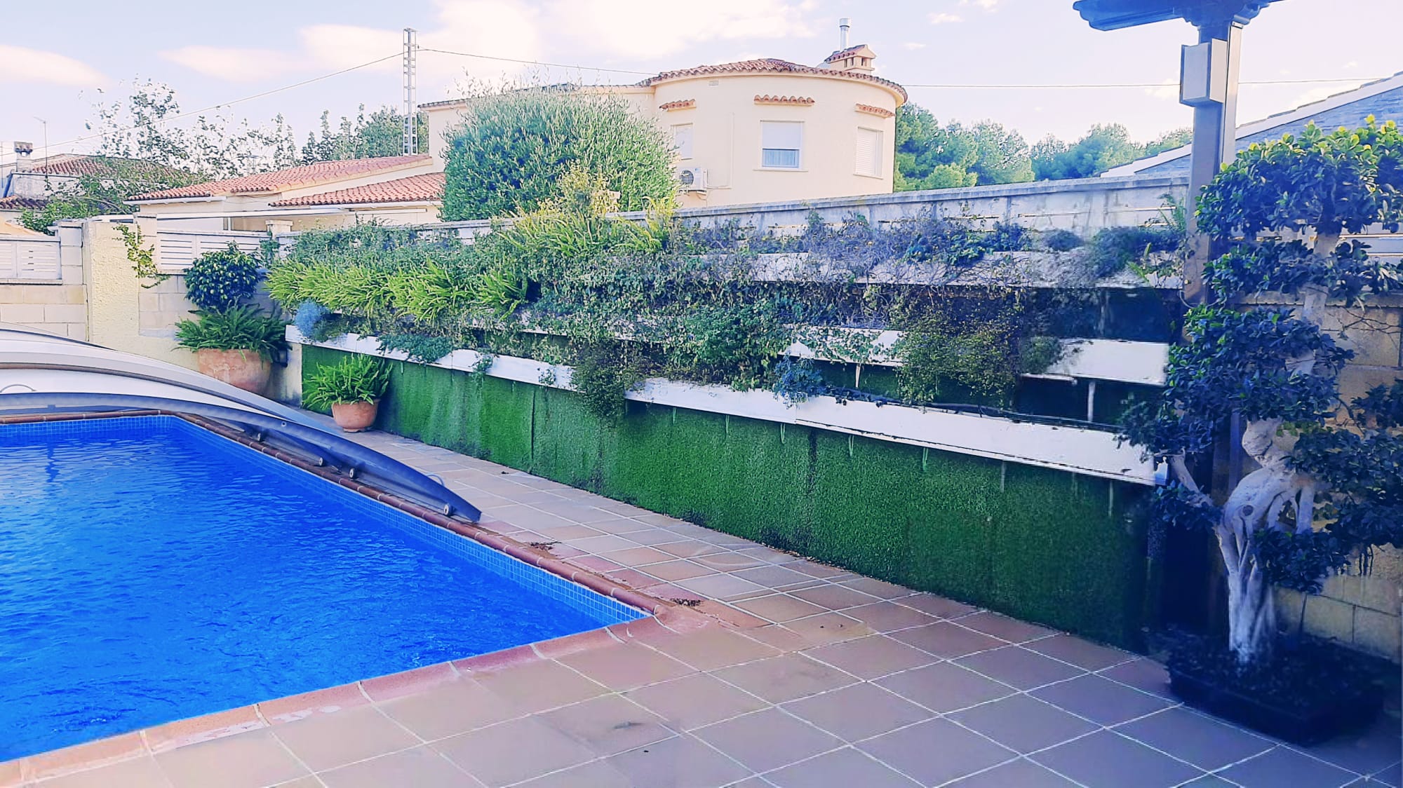 Zona de piscina exterior con terraza de baldosas, cubierta retráctil y un jardín vertical escalonado sobre un paramento de césped artificial.