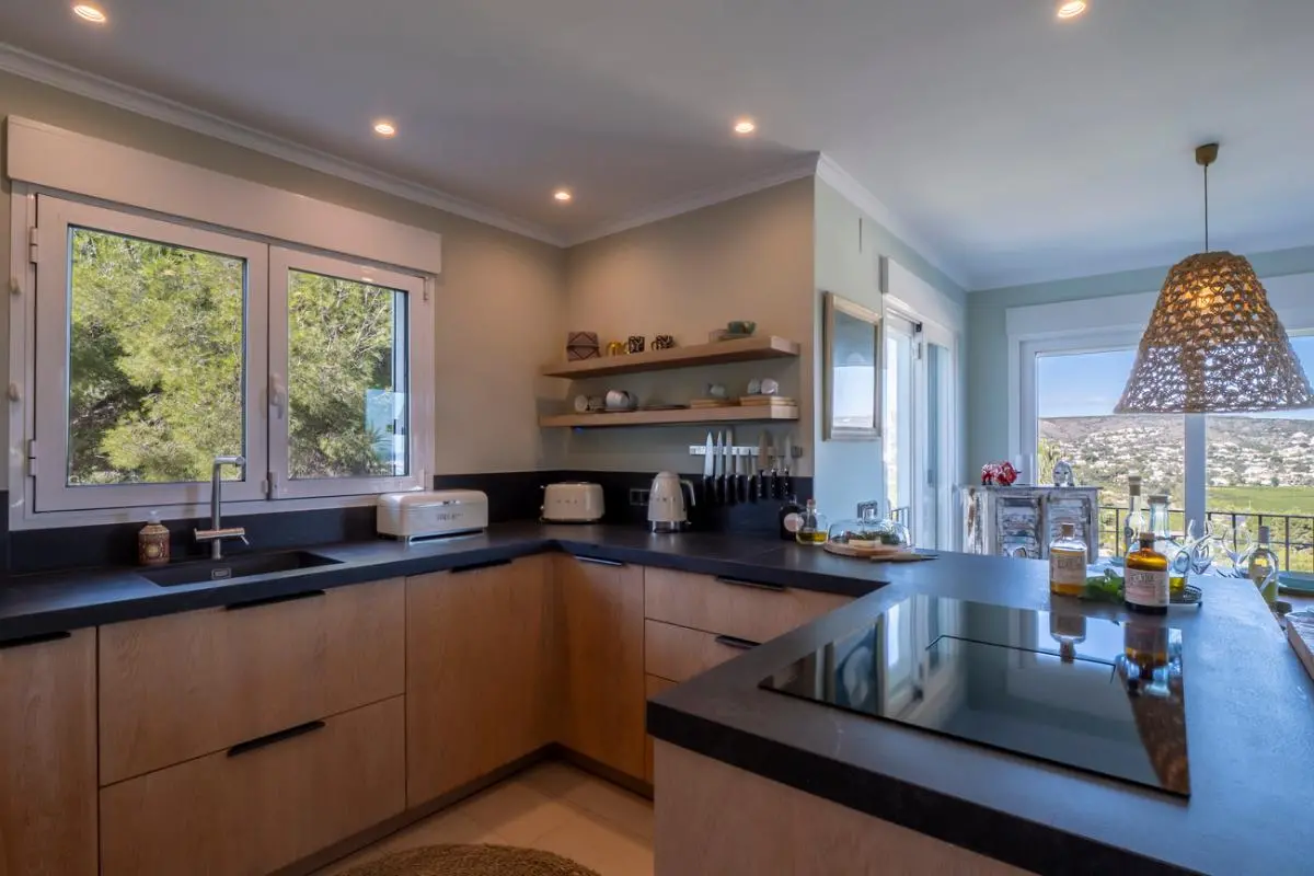 Cocina moderna con armarios de madera clara, encimeras oscuras y placa de inducción. Los amplios ventanales ofrecen luz natural y vistas a los pinos y al valle circundante.