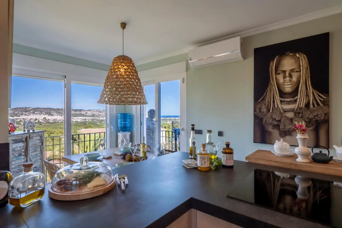 Cocina moderna con encimera oscura y placa de inducción, zona de comedor con lámpara de mimbre y vistas panorámicas a la costa a través de ventanales.
