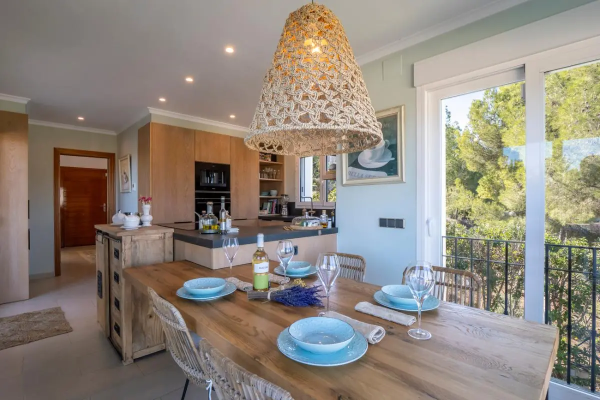 Comedor integrado con mesa de madera maciza, lámpara de fibras naturales y cocina moderna con armarios de roble. Acceso directo a terraza con vistas arboladas.