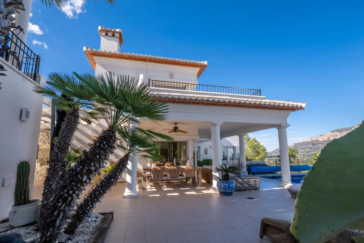 Exterior de una villa blanca con porche de columnas, comedor al aire libre y jardín mediterráneo con vistas despejadas a las montañas.