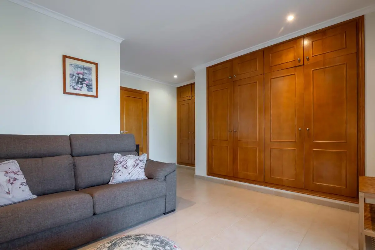 Habitación con un amplio armario empotrado de madera, sofá tapizado en gris y suelos de gres claros iluminados por focos empotrados.