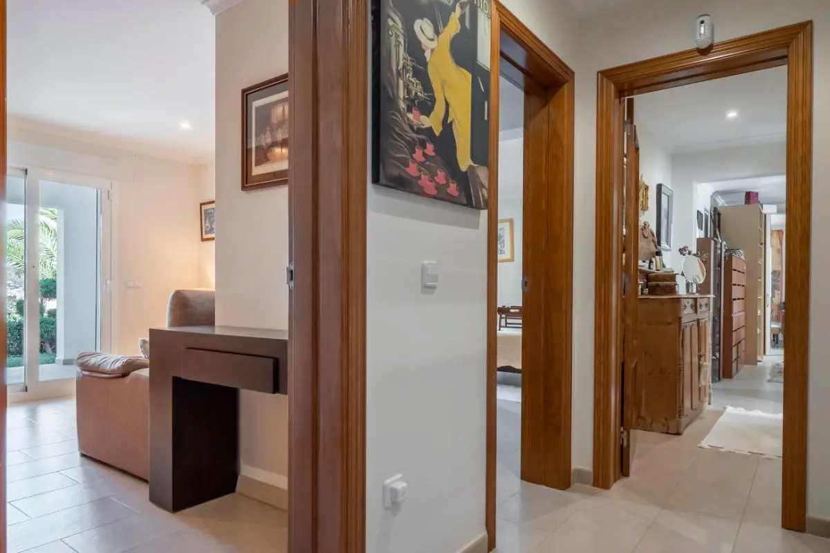 Pasillo con marcos de madera natural, suelos cerámicos claros y acceso visual al salón y habitaciones contiguas con mobiliario de estilo clásico.