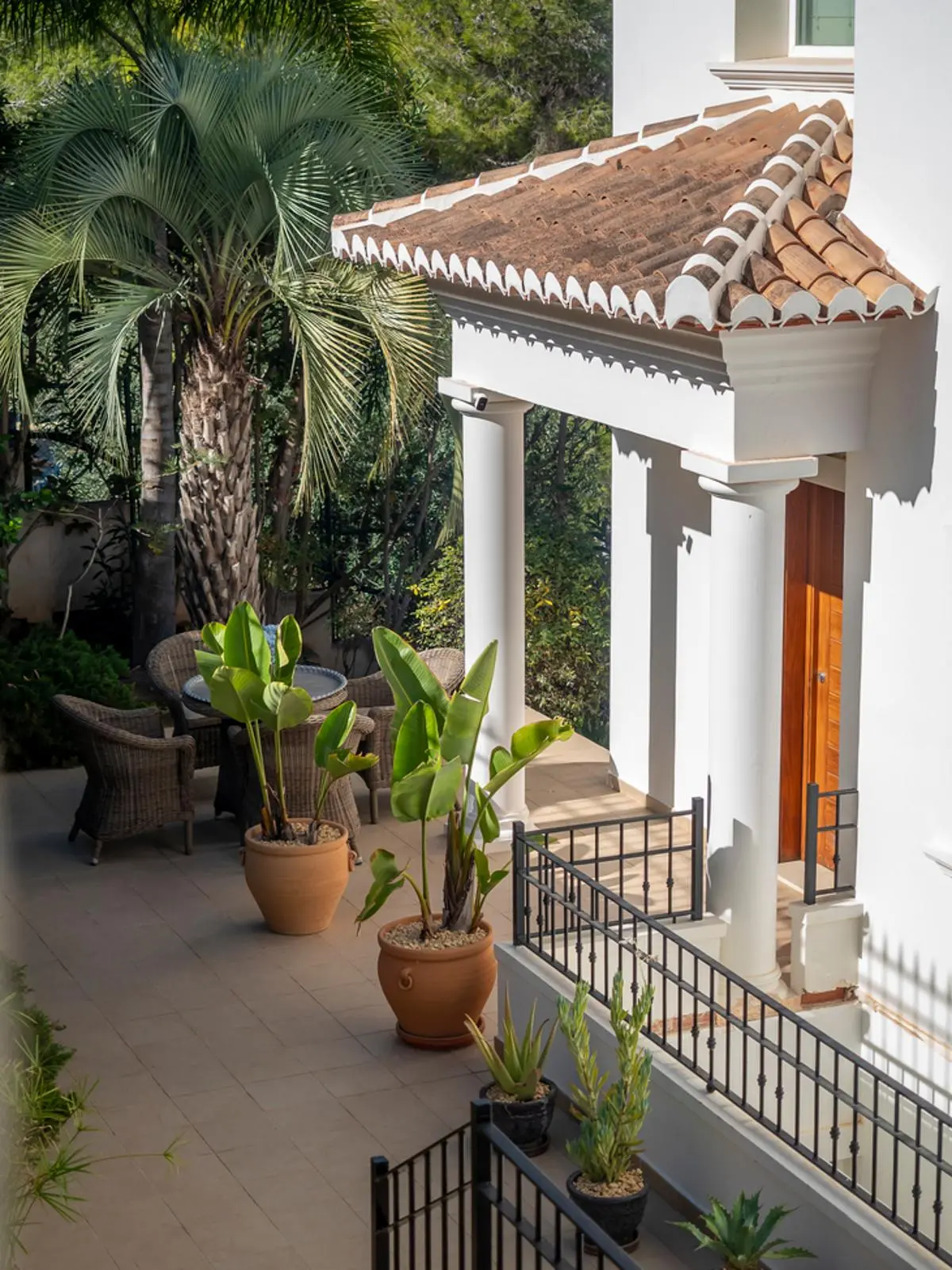 Porche de entrada con columnas blancas y tejado de teja. El patio pavimentado cuenta con plantas tropicales en macetas, barandillas de forja y una puerta de madera.