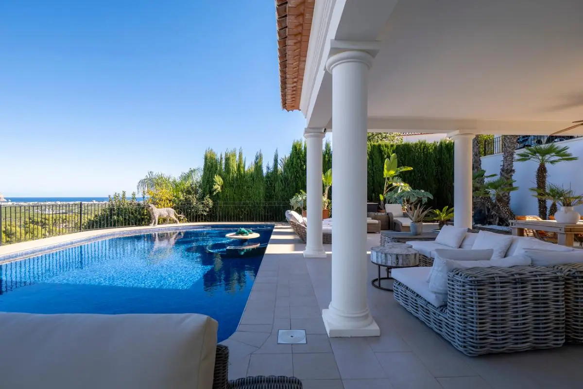 Terraza cubierta con columnas clásicas blancas y sofás de mimbre, situada junto a una piscina de gresite con vistas despejadas al mar.