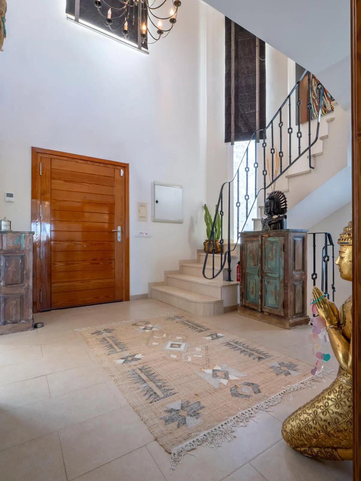 Vestíbulo con puerta de madera maciza, escalera de piedra con barandilla de hierro forjado y techos altos. Incluye mobiliario rústico y suelos de baldosa clara.