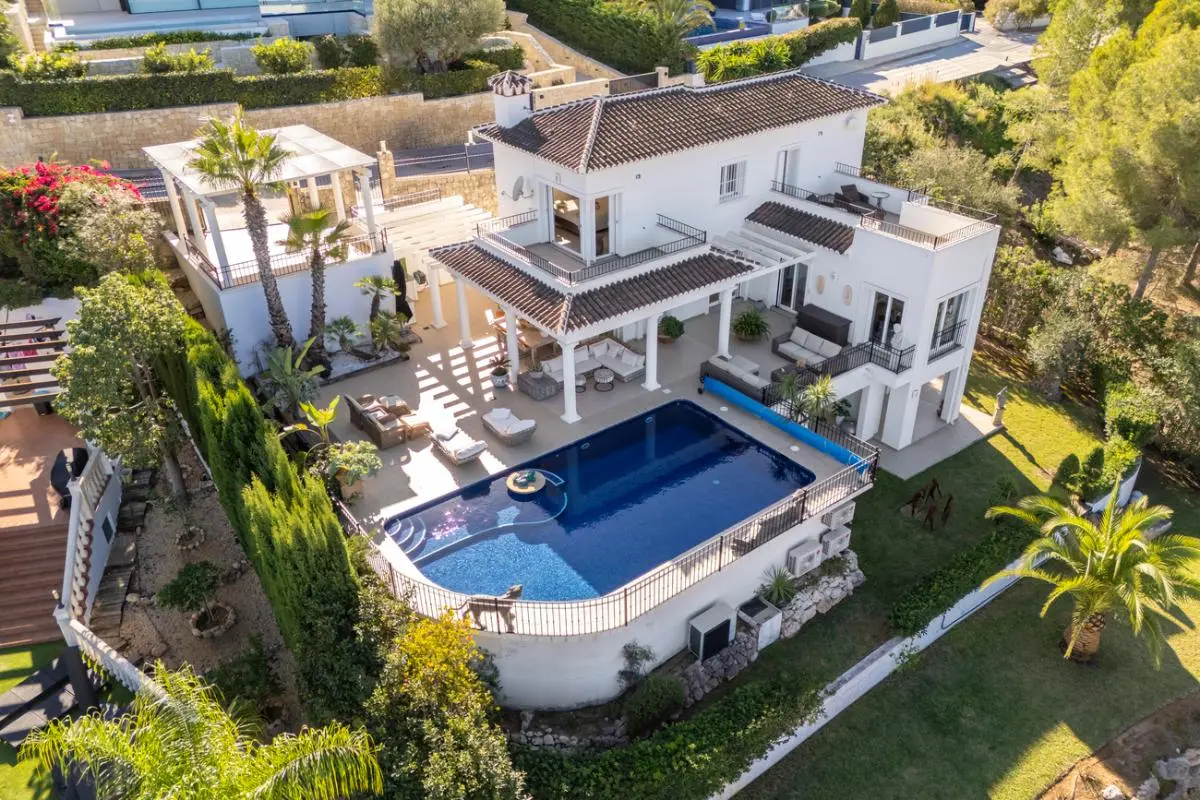 Vista aérea de una villa mediterránea blanca con piscina, múltiples terrazas cubiertas con columnas y jardines paisajísticos con palmeras.