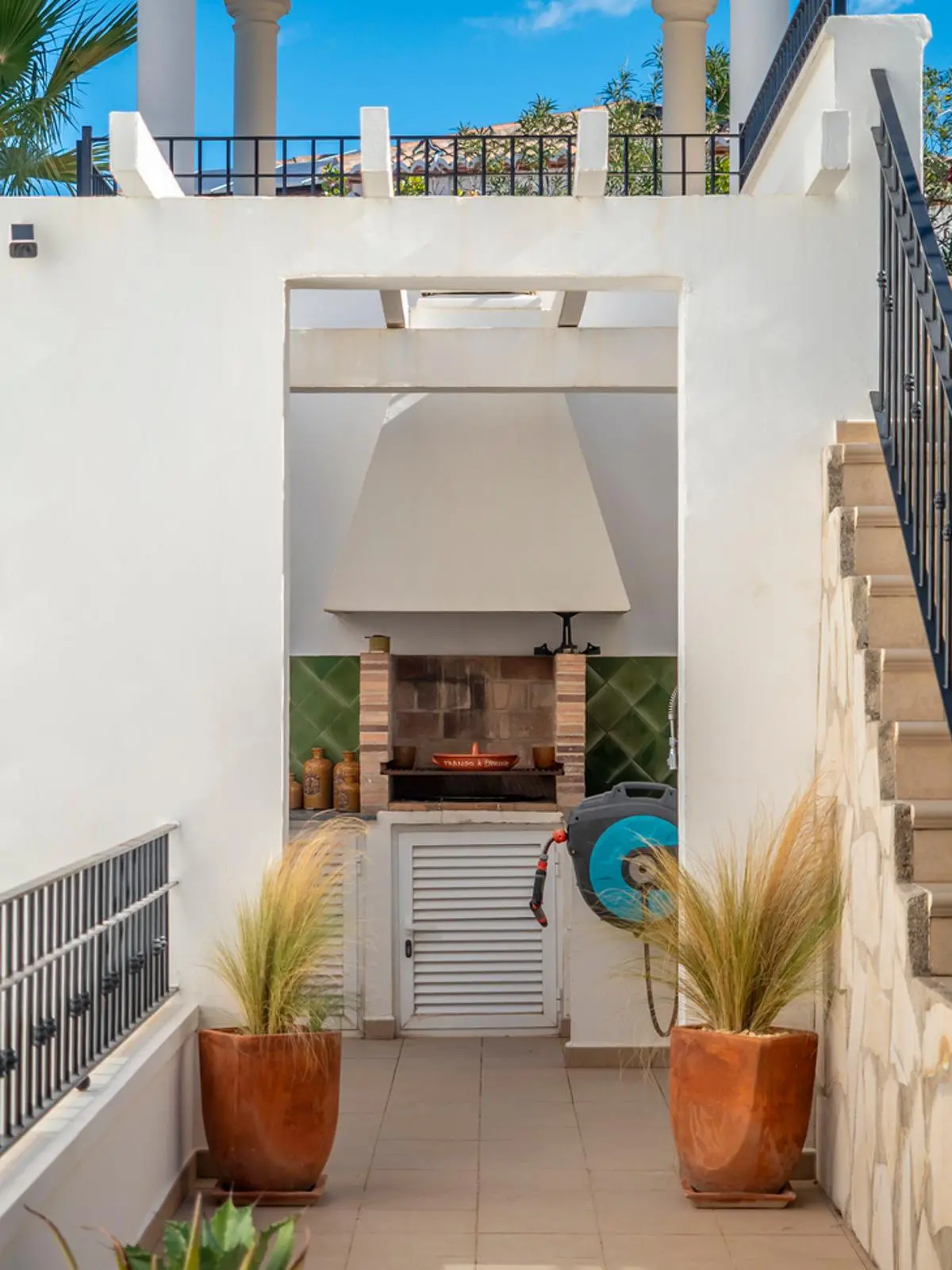 Zona de barbacoa exterior con parrilla de ladrillo, campana blanca y salpicadero de azulejos verdes, flanqueada por maceteros de terracota y paredes blancas.