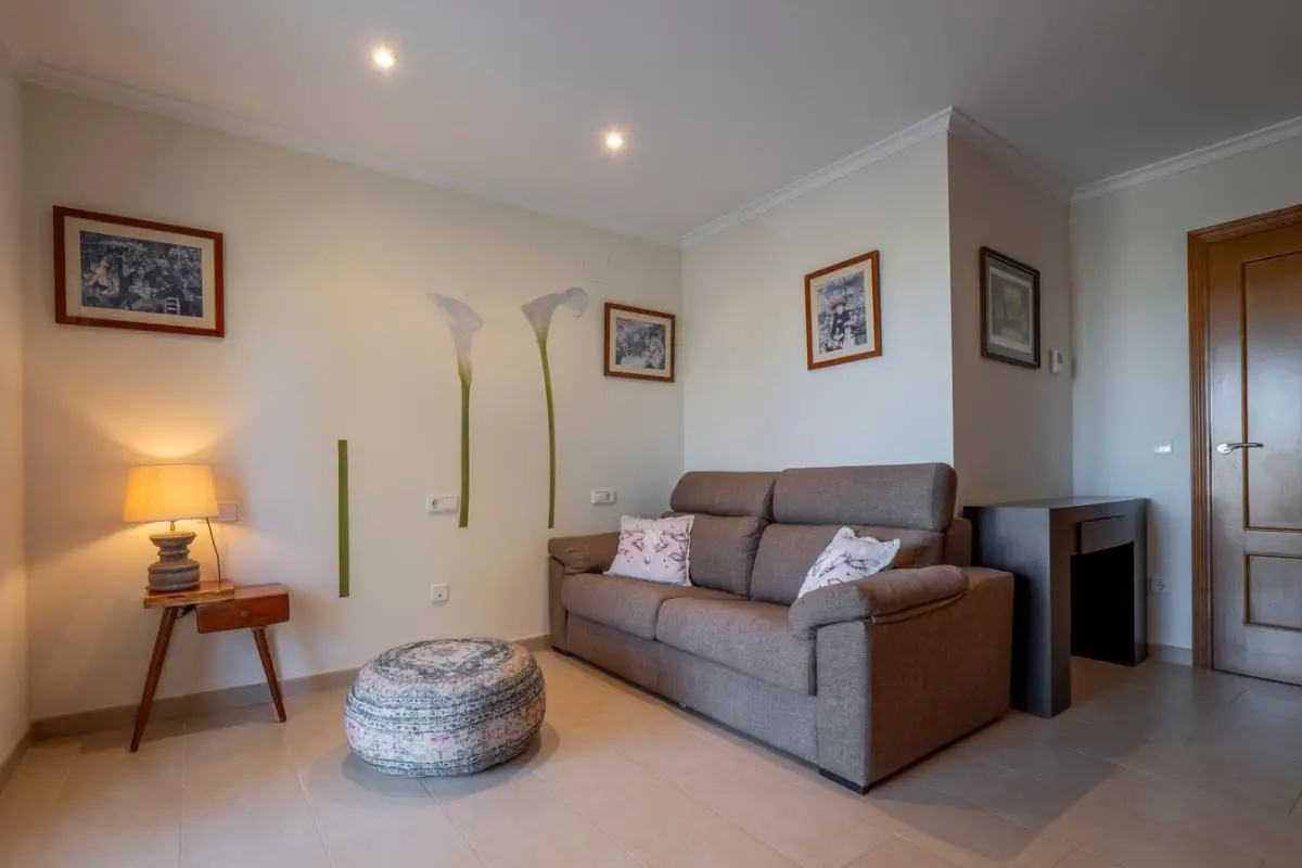 Zona de estar con sofá de tela marrón, puf estampado y mesa auxiliar de madera. La pared incluye vinilos florales, cuadros enmarcados e iluminación empotrada.
