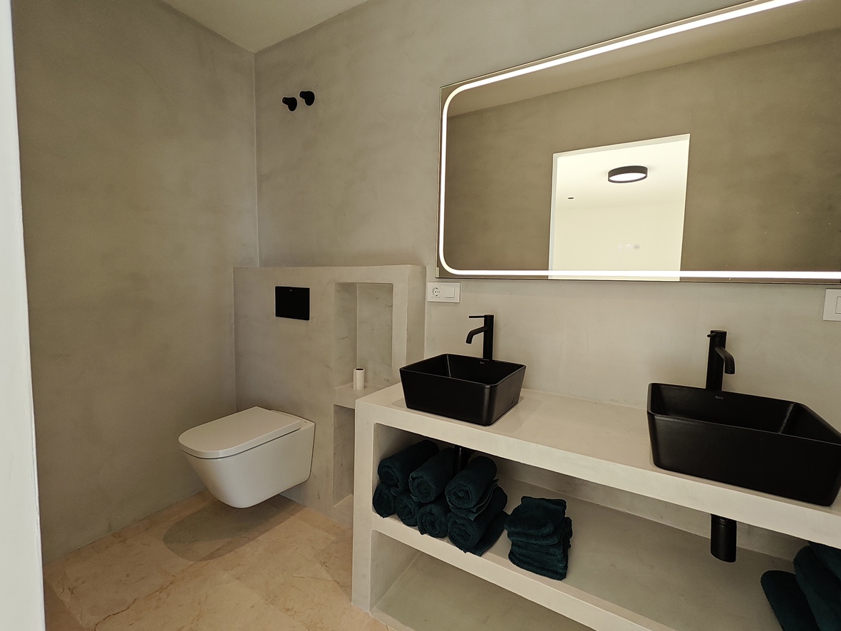 Baño de diseño minimalista con paredes de microcemento gris, lavabo doble con senos negros y un amplio espejo con iluminación perimetral integrada.