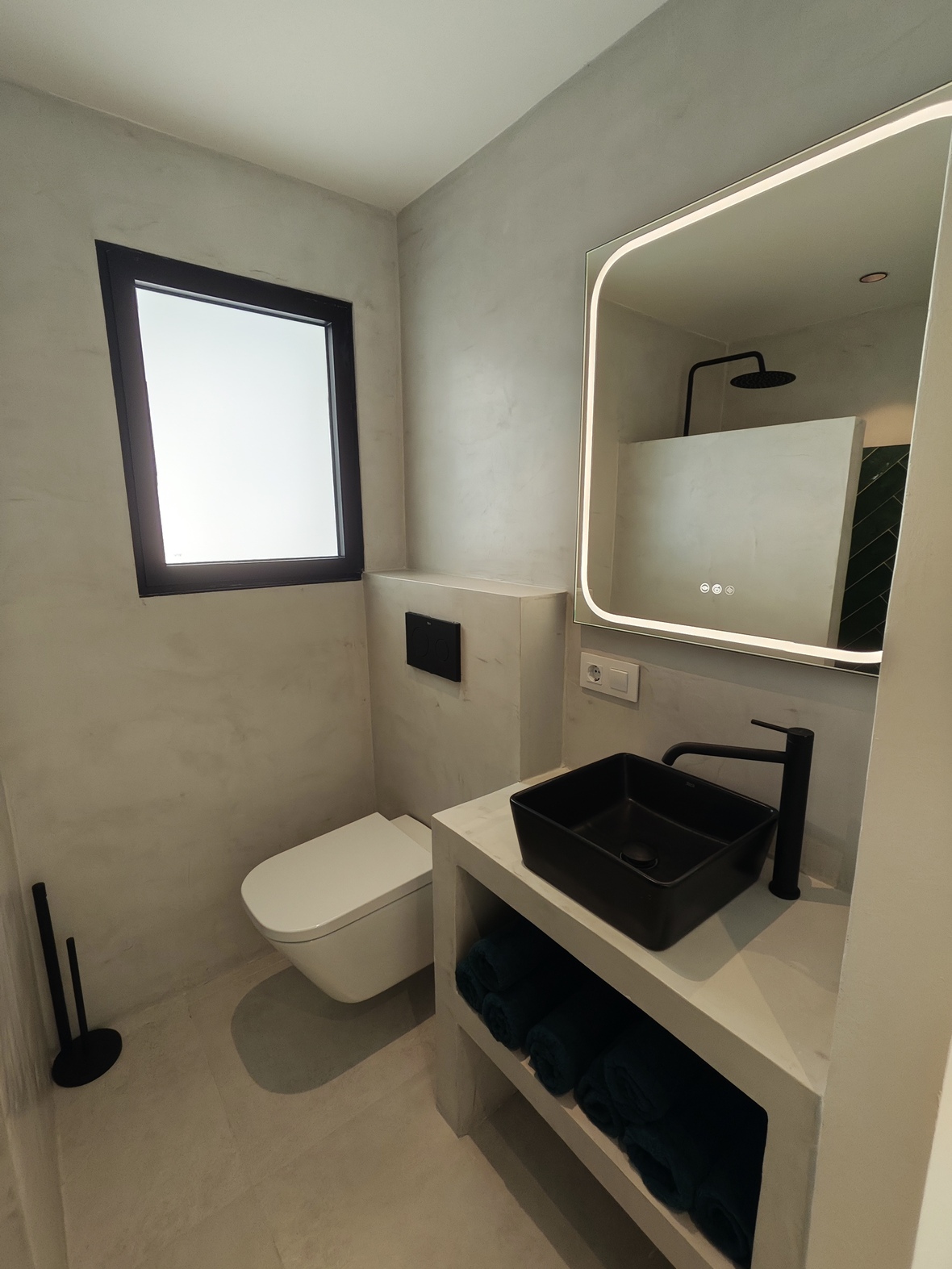 Baño moderno con acabados de microcemento, lavabo de sobremesa negro mate y espejo retroiluminado. Incluye grifería negra y un inodoro suspendido de diseño.