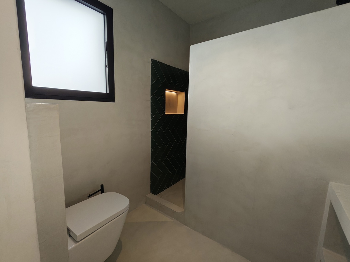 Baño moderno con paredes de microcemento, inodoro suspendido y ducha de obra con azulejos verdes en espiga y una hornacina iluminada.