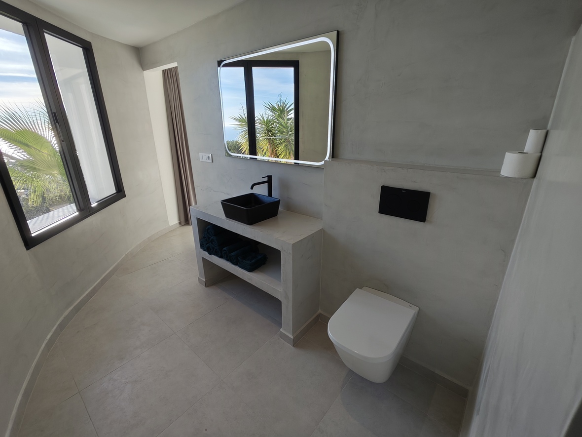 Baño moderno con paredes de microcemento, lavabo negro sobre encimera de piedra y espejo retroiluminado. Amplios ventanales ofrecen luz natural y vistas exteriores.
