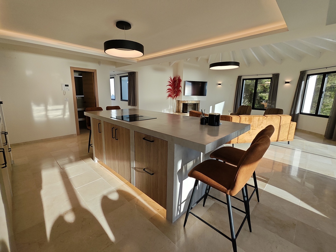 Cocina de concepto abierto con isla de acabado cemento, placa de inducción y taburetes de cuero, bajo un techo de vigas blancas y luminarias circulares modernas.