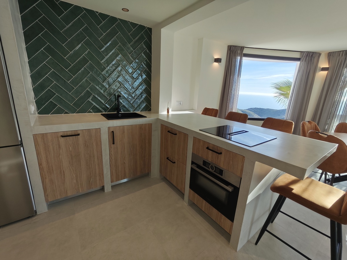 Cocina moderna con salpicadero de azulejos verdes en espiga, armarios de madera y una isla con placa de inducción y vistas panorámicas al mar.