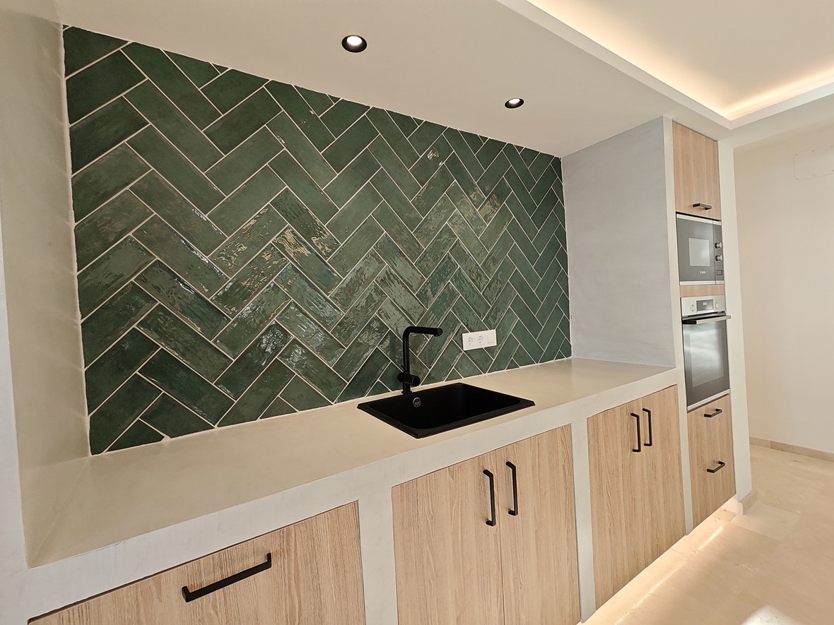 Cocina moderna con salpicadero de azulejos verdes en espiga, fregadero negro y armarios de madera clara con electrodomésticos integrados e iluminación LED.