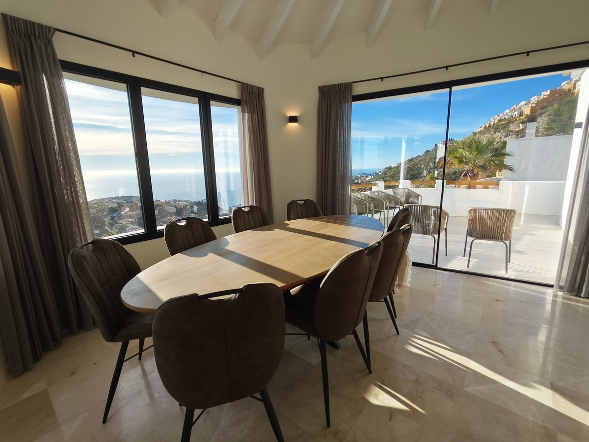 Comedor con mesa ovalada de madera y suelo de mármol, con amplios ventanales que ofrecen vistas panorámicas al mar y acceso directo a la terraza exterior.