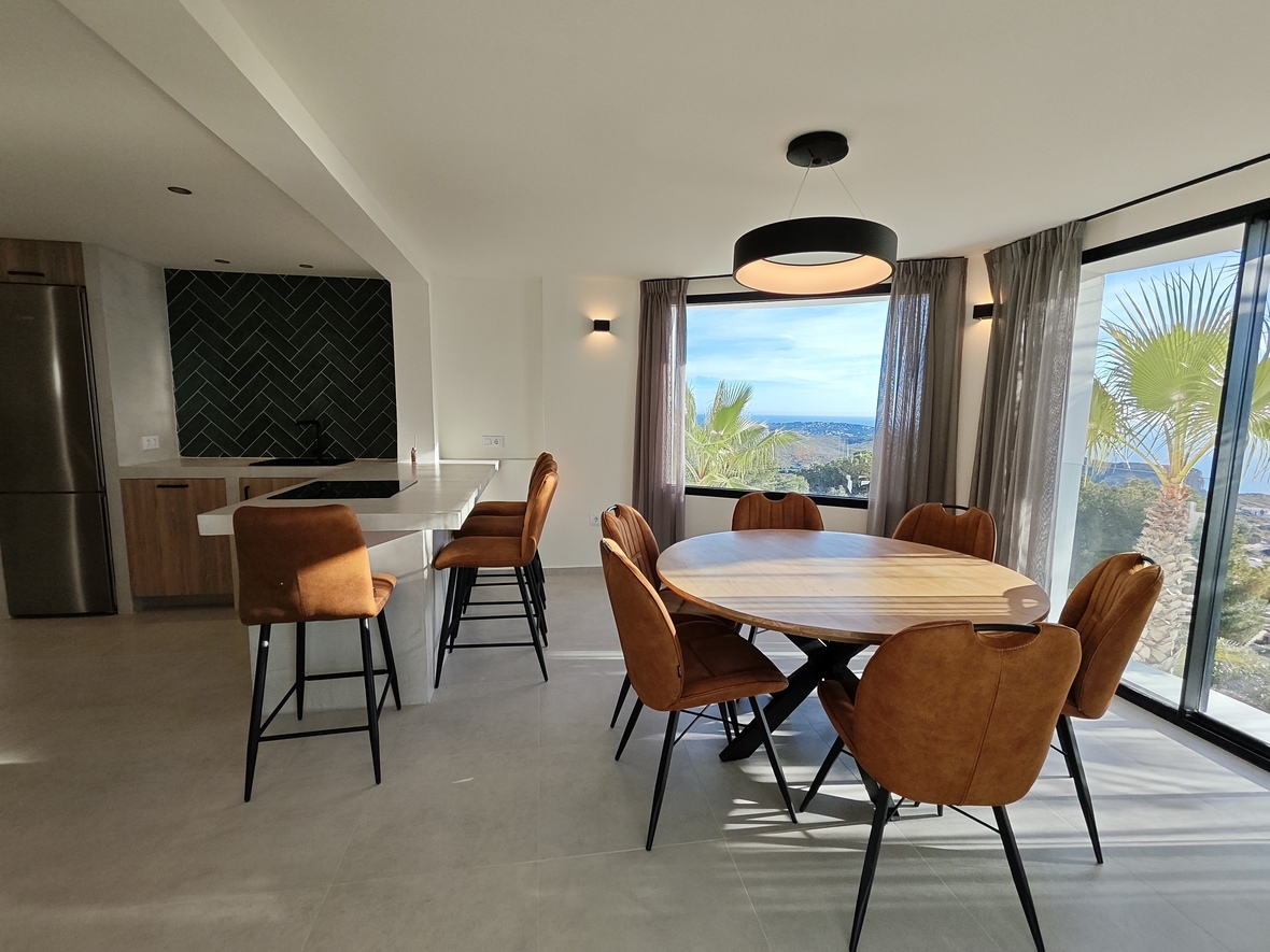 Comedor de planta abierta con mesa circular de madera, sillas color coñac y cocina de diseño con salpicadero de espiga y vistas panorámicas al mar.