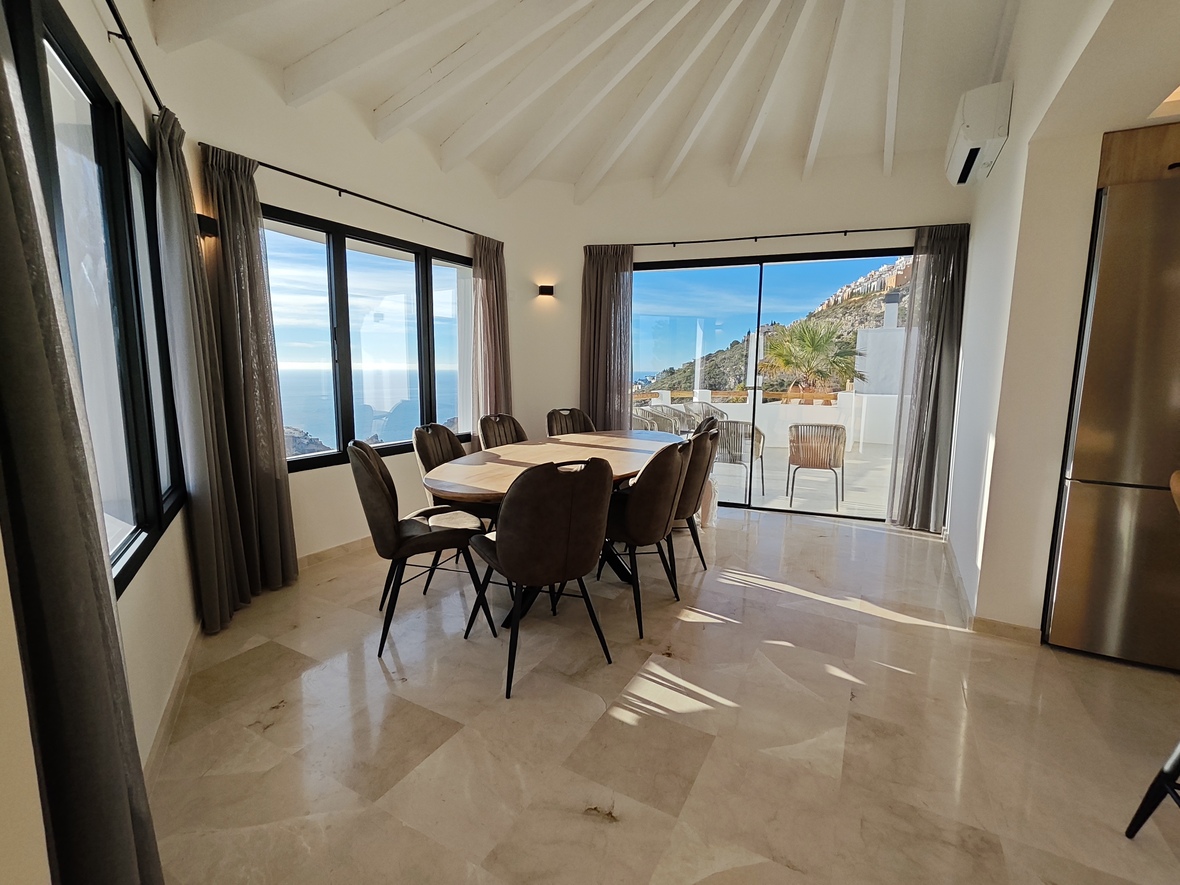 Comedor luminoso con mesa ovalada de madera, suelo de mármol y techo abovedado con vigas. Los ventanales ofrecen vistas al mar y acceso directo a la terraza.