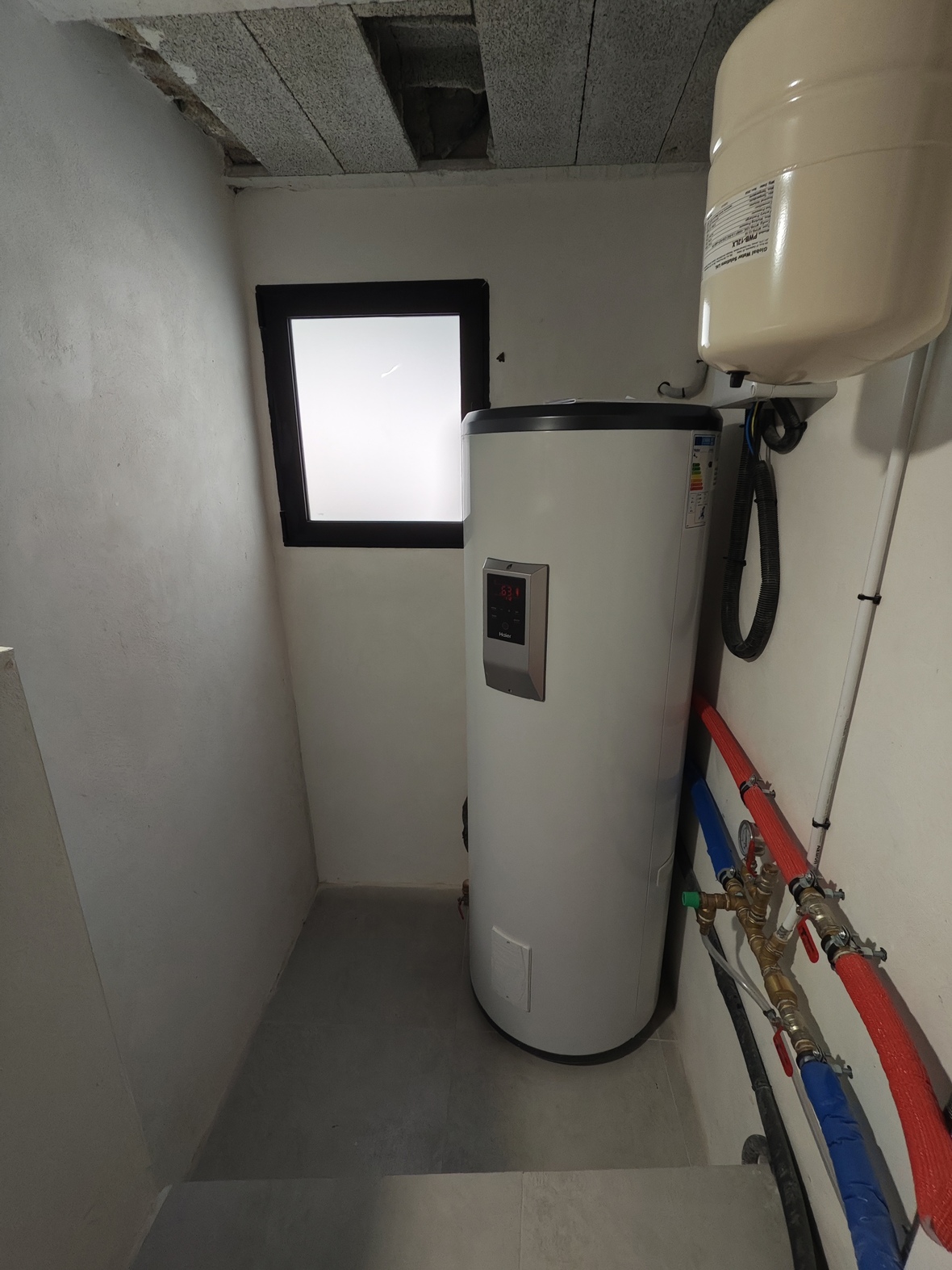 Cuarto de instalaciones con termo aerotérmico Haier, vaso de expansión y tuberías aisladas. Incluye una ventana de luz natural y suelo de gres gris.