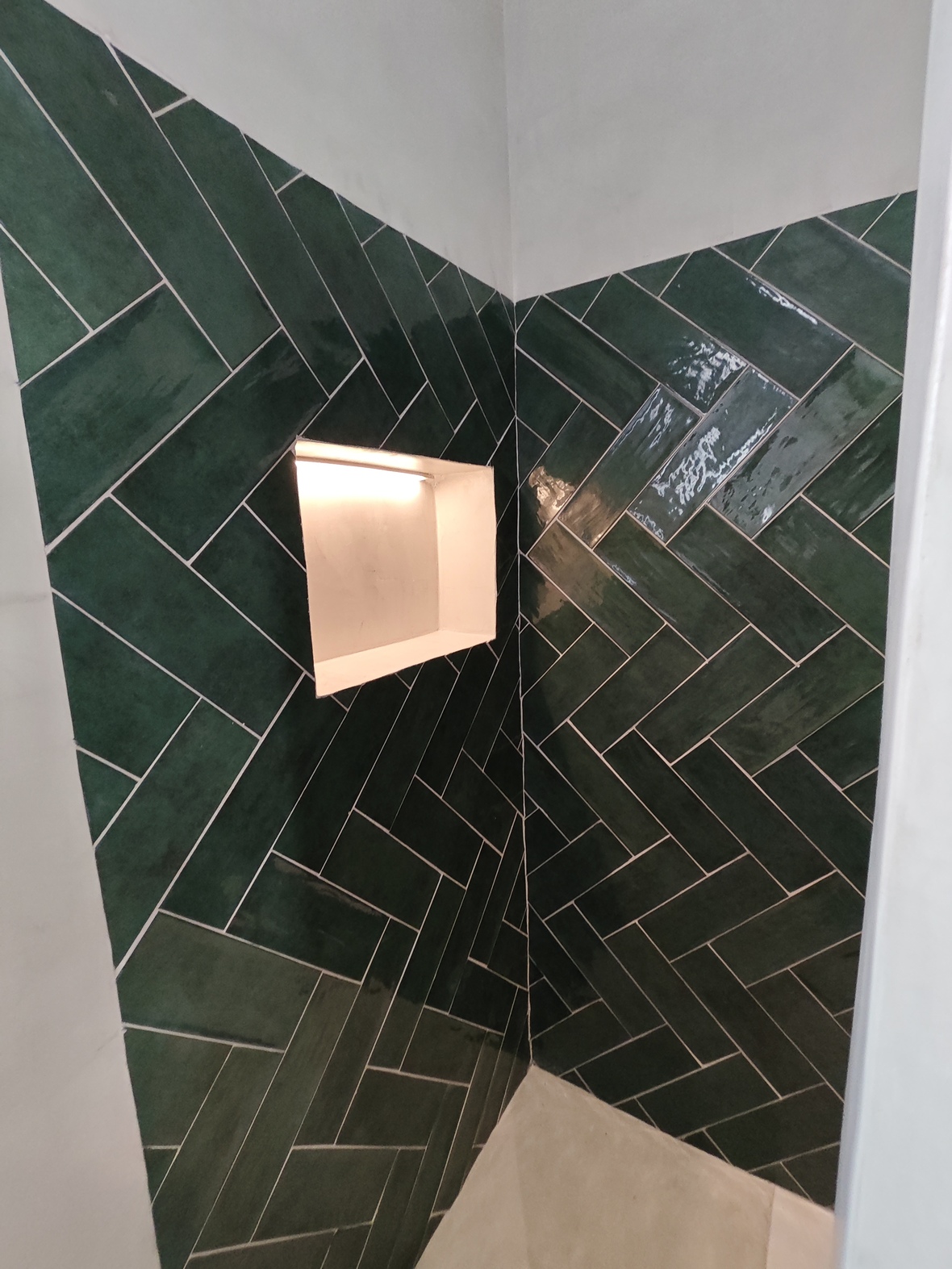 Detalle de ducha con azulejos verde esmeralda dispuestos en espiga y una hornacina empotrada con iluminación LED cálida integrada.