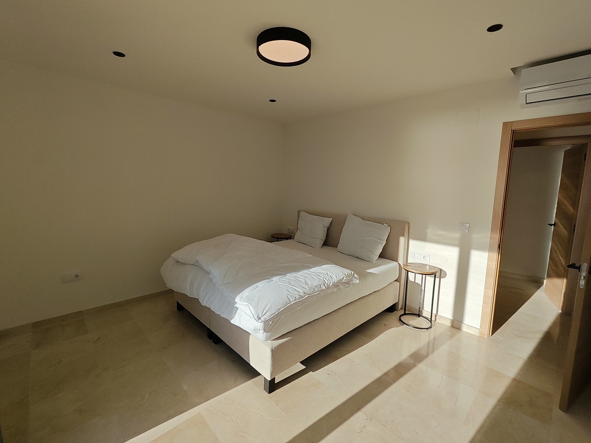 Dormitorio minimalista con suelo de piedra pulida, cama doble tapizada e iluminación moderna que incluye un plafón circular y focos empotrados.