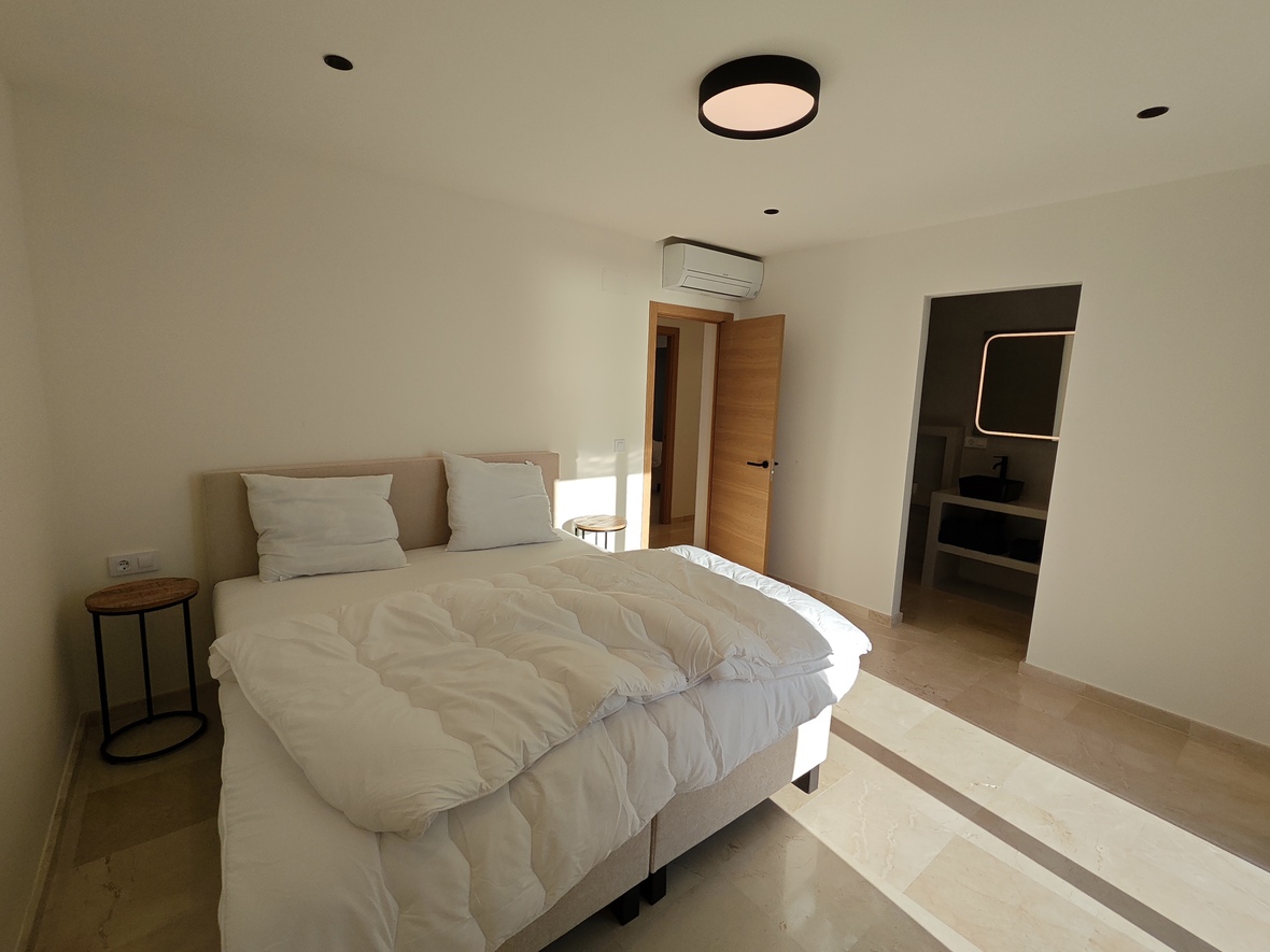 Dormitorio moderno con suelos de piedra, cabecero tapizado en beige y mesitas minimalistas. Incluye aire acondicionado y acceso a un baño en suite con espejo retroiluminado.