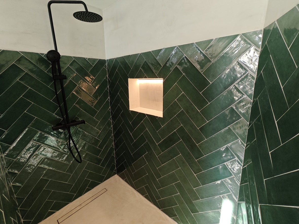 Ducha con azulejos verdes brillantes en espiga, grifería negra mate y hornacina iluminada con LED. Suelo de resina clara con desagüe lineal integrado.