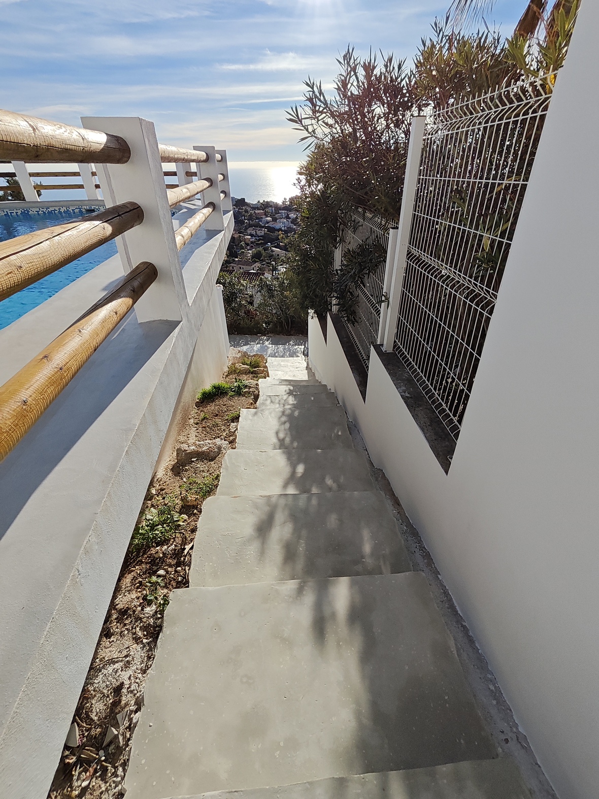 Escalera exterior de hormigón con barandillas de madera y muros blancos, con vistas despejadas hacia la costa mediterránea y la zona de piscina.